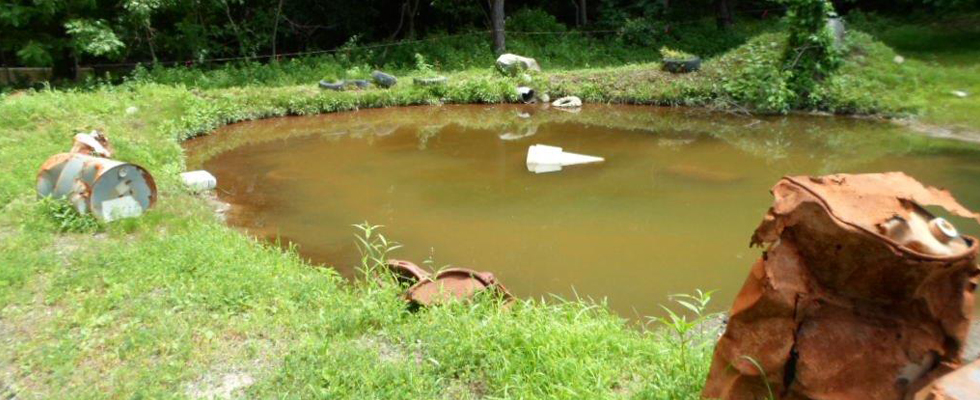 Photo of cloudy water in a small holding pond located in front of a densly wooded area. Rusted 55-gallon drums are located on the ground adjacent to the pond.