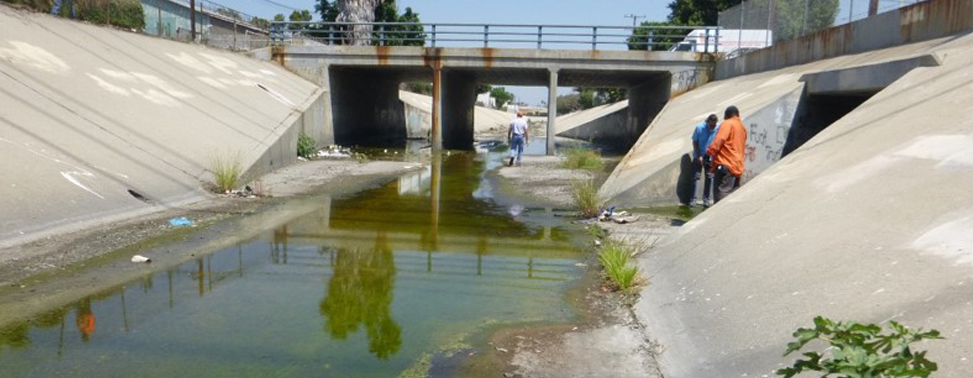 Photo of inspectors walking in a concrete channelized stream with minimal flow. A small road passes over the channel via a culvert.